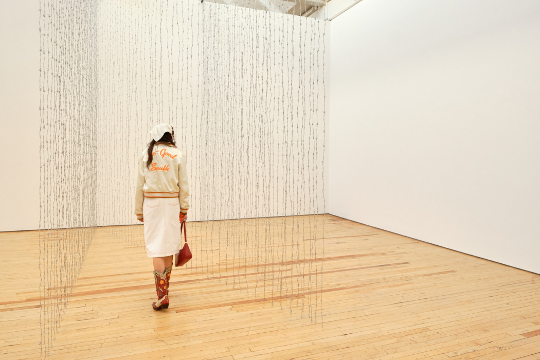 A woman walks between a sculpture made of barb wire that runs in a diagonal lines.